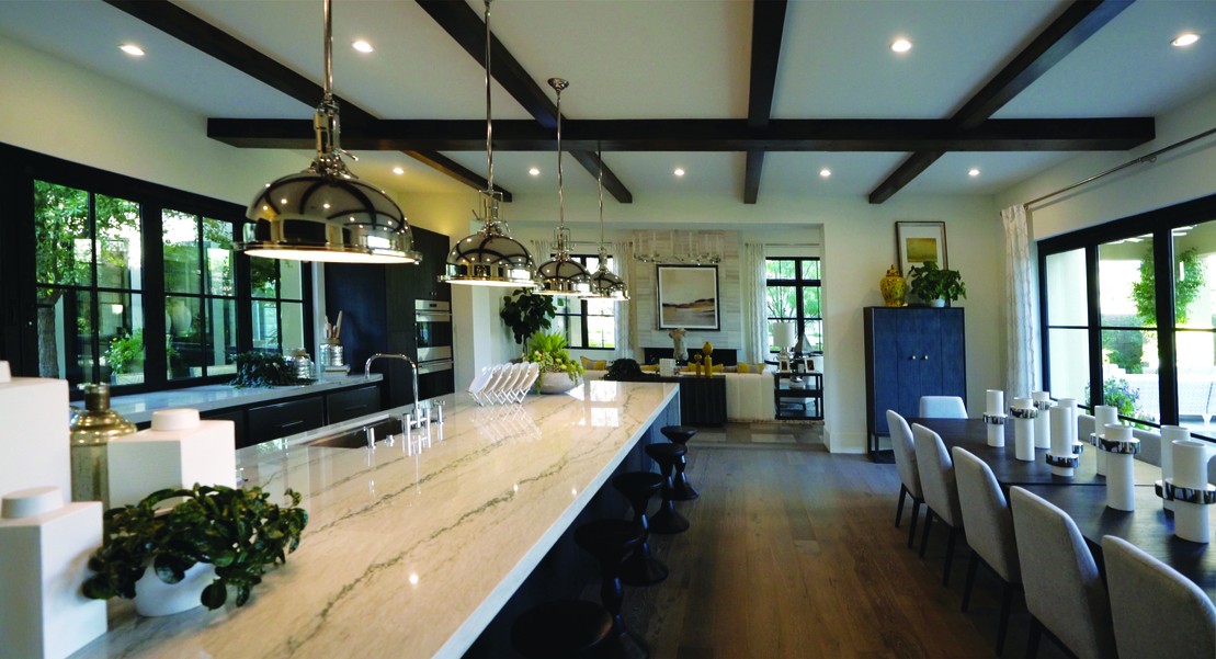 A spacious modern kitchen and dining area featuring a long marble island with black barstools, sleek black cabinetry, pendant lighting, exposed ceiling beams, and large windows.